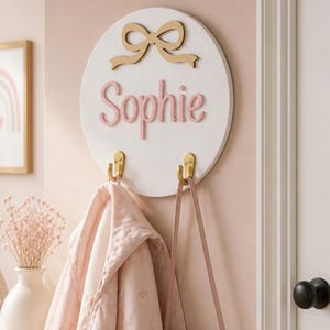 Peut inclure: Un porte-manteau mural circulaire en bois blanc avec une découpe de nœud doré sur le dessus et le prénom Sophie en lettres cursives roses en relief. Deux crochets en métal doré sont fixés en bas pour suspendre des objets.