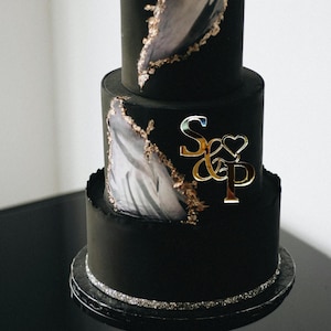 May include: A three-tiered black cake with gold accents and a silver base. The cake features a gold "S & P" monogram and abstract gray and gold designs. The cake is likely for a wedding or special event.