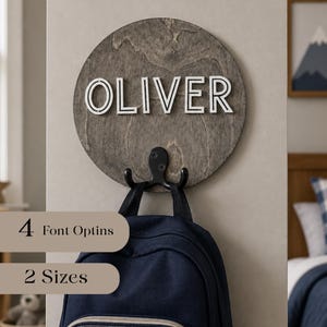 Peut inclure: Une plaque murale circulaire en bois avec une finition teintée gris foncé présentant le nom OLIVER en relief blanc. Un crochet double en métal noir est fixé en bas, supportant un sac à dos bleu marine. Le texte indique 4 Font Options et 2 Sizes.