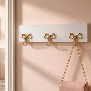 Peut inclure: Une étagère murale rectangulaire blanche dotée de trois crochets en métal doré en forme de nœuds. Chaque crochet possède une base incurvée pour suspendre des objets. Un sac à main rose clair avec une bandoulière en chaîne dorée est suspendu.
