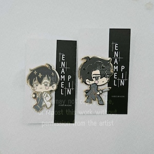 Puede incluir: Dos pines de esmalte con personajes de estilo anime. Uno muestra un personaje con un atuendo blanco y negro, el otro con un atuendo gris sosteniendo un cuchillo. Cada pin está en una tarjeta negra con el texto "ENAMEL PIN".