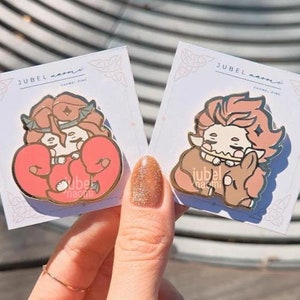 May include: Two enamel pins featuring cartoon characters. The first pin is red and features a character with red hair and a crown. The second pin is pink and features a character with orange hair and a crown. Both pins have the text "JUBEL" on them.