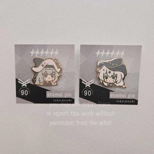 May include: Two enamel pins featuring a cartoon character with a hat. The pins are on gray cards with the number 90 and the text "enamel pin" and "JUBELNAOMI". The pins have a gold outline and are in a cartoon style.