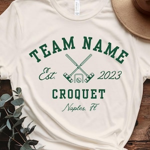 May include: Cream-colored t-shirt with the text "TEAM NAME" in green, arched above "CROQUET" and "Naples, Fl." The design includes crossed mallets and the year "2023". A casual, sporty apparel item.