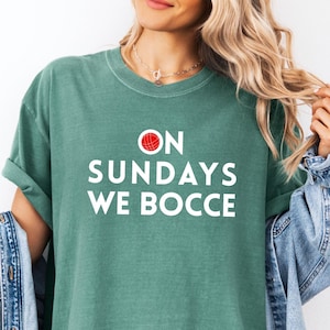 May include: A sage green short-sleeve t-shirt featuring the white text ON SUNDAYS WE BOCCE, with a small red circular graphic replacing the letter O. The shirt is worn with a light blue denim jacket draped over the shoulders.