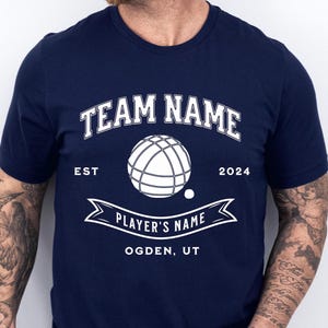 Peut inclure: Un t-shirt bleu marine avec un motif blanc. Le motif représente une boule de pétanque blanche, le texte "TEAM NAME EST 2024", une bannière avec le texte "PLAYER'S NAME", et le texte "OGDEN, UT".