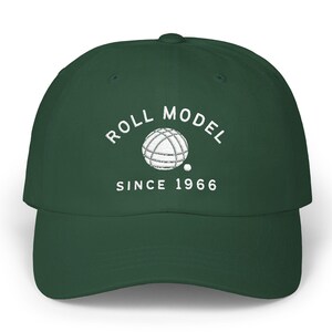 Peut inclure: Une casquette de baseball vert foncé avec une broderie blanche indiquant ROLL MODEL au-dessus d'un dessin de boule de pétanque et d'un petit cochonnet, avec le texte SINCE 1966 brodé en blanc sous le motif sur le panneau avant de la casquette.