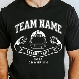 May include: Black t-shirt with a white graphic design featuring a football helmet, footballs, and the text "TEAM NAME" above "LEAGUE NAME" and "2026 CHAMPION". The design is centered on the shirt.