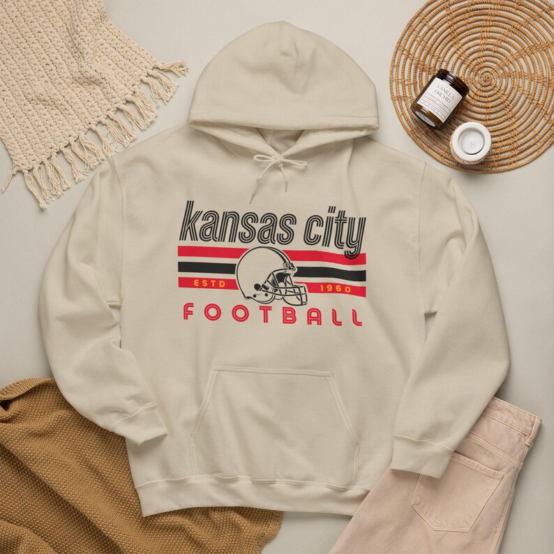 May include: A cream-colored hooded sweatshirt featuring a graphic print that reads "kansas city" in black, followed by red and black horizontal stripes, a football helmet illustration, "ESTD 1960" in red, and "FOOTBALL" in red lettering.