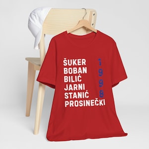May include: A red t-shirt with white text listing names and the number 1998 in blue. A white baseball cap rests on a wooden chair behind the shirt.