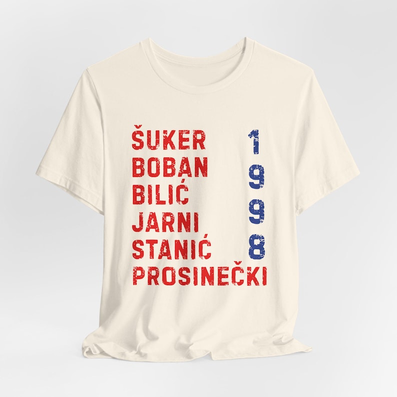 May include: Cream-colored t-shirt with red and blue text. The shirt features a list of names in red and the number "1998" in blue. The text has a distressed, vintage look.