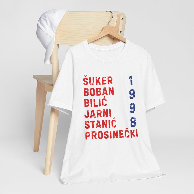 May include: White t-shirt with red and blue text. The shirt displays names and the number 1998. A white baseball cap rests on a wooden chair behind the shirt.