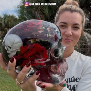 Könnte beinhalten: Ein durchsichtiger Schädel aus Harz, gefüllt mit getrockneten roten Rosen und anderen Blumen. Der Schädel wird von einer Person mit rotem Nagellack gehalten.