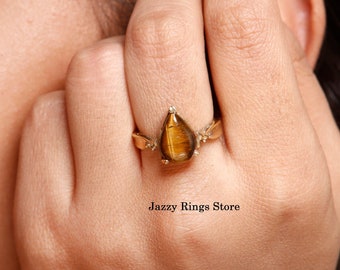 May include: A gold ring with a teardrop-shaped tiger's eye gemstone. The ring is set in a delicate gold band with small accents. "Jazzy Rings Store" is printed below the ring.