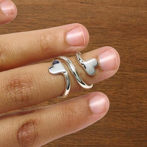 May include: A silver ring with a heart-shaped design. The ring wraps around the finger with two heart-shaped accents. The ring is shown on a finger against a brown wooden background. Jewelry.
