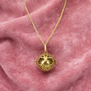 Puede incluir: Un collar dorado con un colgante con un diseño de esfera de filigrana. La esfera tiene un patrón calado con un motivo de árbol de la vida. La cadena del collar es delicada y cuelga sobre un fondo de terciopelo rosa.