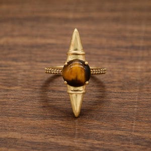 May include: A gold-colored ring featuring a large, round, brown tiger's eye stone. The ring has a unique design with a central vertical spike and decorative elements on the band. The ring is set against a wooden background.