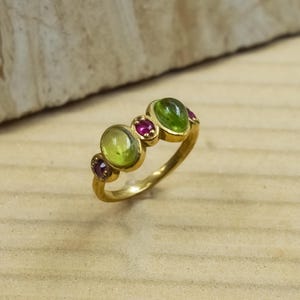 May include: A gold ring featuring three oval green gemstones and two smaller red gemstones. The ring has a classic design with a polished finish, set against a light wooden surface.