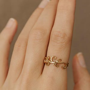 Puede incluir: Un anillo dorado con un intrincado diseño de vid en espiral. El anillo se lleva en un dedo, mostrando los delicados detalles del patrón floral. El diseño del anillo presenta una serie de rizos y hojas, creando una estética elegante e inspirada en la naturaleza.