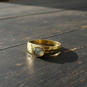 Puede incluir: Un anillo dorado con textura martillada y una piedra ovalada de color claro. El anillo está sobre una superficie de madera oscura, con la sombra de la luz solar.