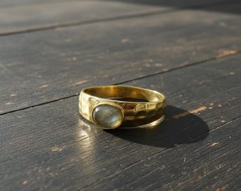 May include: A gold-colored ring with a hammered texture and an oval, light-colored stone. The ring is set on a dark wooden surface, with the sunlight casting a shadow.