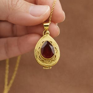 May include: A gold-colored pendant necklace with a teardrop-shaped locket. The locket features a large, faceted, deep red gemstone. The pendant is suspended from a delicate gold chain. The locket has a decorative, textured border.