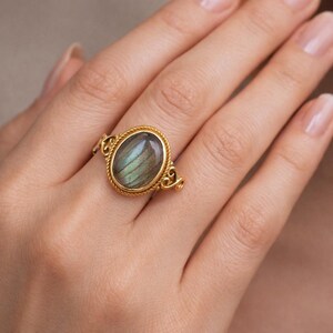 May include: A gold ring with an oval, iridescent gemstone. The ring has an ornate gold setting with a twisted rope design around the gemstone and decorative flourishes. The gemstone displays shades of green and blue. The ring is a piece of jewellery.
