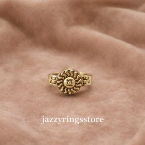 May include: A gold-colored ring with a sun design. The ring features a detailed sunburst pattern around a central sun face. The band has decorative elements. The text "jazzyringsstore" is at the bottom of the image.