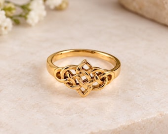 May include: A polished gold ring featuring an intricate, symmetrical Celtic knot design at the centre. The band is smooth and circular, leading into the detailed, interwoven metalwork that forms the focal point of this jewellery piece.