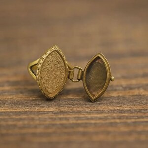 Puede incluir: Un anillo con medallón de color dorado con un compartimento de forma ovalada que se abre. El anillo tiene una superficie texturizada y un borde decorativo alrededor del compartimento. El medallón está conectado por una pequeña bisagra.
