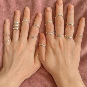 Puede incluir: Dos manos que muestran varios anillos de plata. Los anillos incluyen bandas simples, anillos apilados y anillos con diseños geométricos únicos. Los anillos se presentan sobre un fondo rosa.