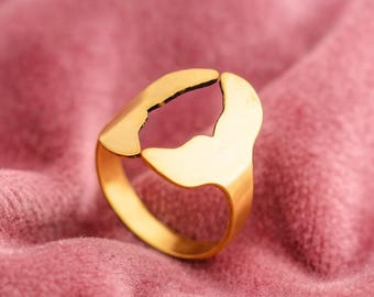 May include: A gold-colored ring with a unique, open design. The ring features a smooth, polished finish and a distinctive shape with a cutout detail. The ring is set against a pink, textured background.