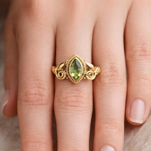 May include: A gold ring featuring a marquise-cut, light green gemstone. The ring has an ornate, filigree design with a twisted rope border around the stone. The band has decorative swirls on each side.