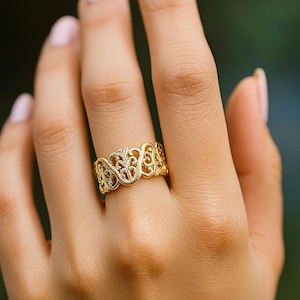 Puede incluir: Un anillo dorado con un intrincado diseño calado. El anillo presenta un patrón repetitivo de formas arremolinadas y elementos florales. El anillo se lleva en un dedo, mostrando su detallada artesanía y su elegante estilo.