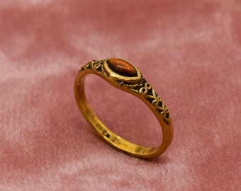 May include: A gold-colored ring with an oval, brown gemstone. The ring has an ornate design around the gemstone and along the band. The ring is set against a pink background.