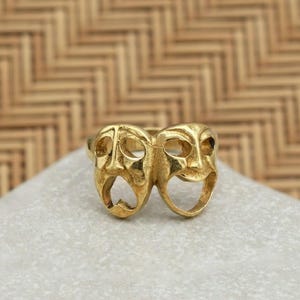 May include: A gold-colored ring featuring the classic comedy and tragedy theater masks. The ring is set on a white surface, with a woven, light brown background. The masks are detailed with open eyes and mouths, and the ring appears to be made of metal.