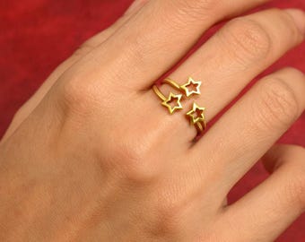 May include: A gold-coloured ring featuring three star-shaped cutouts. The ring is worn on a finger, with the stars arranged in a cluster. The background is a solid red colour.