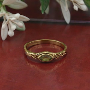 Peut inclure: Une bague dorée avec une pierre précieuse ovale vert clair. La monture présente un motif filigrané orné. La bague est posée sur un fond rouge foncé, avec des fleurs blanches dans le coin supérieur gauche.