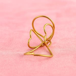 May include: A gold-colored metal sculpture with a heart-shaped design. The sculpture has a looped top and a triangular base. The background is a solid pink color.