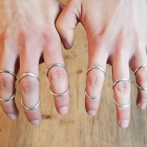 May include: Two hands adorned with multiple silver rings. The rings vary in design, some fully encircling the fingers, others partially. The jewellery is set against a wooden surface, contrasting with the metallic shine.