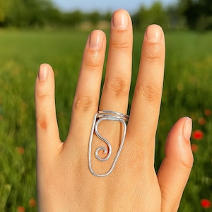 Puede incluir: Un anillo de alambre plateado con un diseño abstracto único. El anillo presenta un marco rectangular con una forma arremolinada, similar a la cursiva, en el interior. El anillo se lleva en una mano, con un campo verde y cielo azul de fondo.