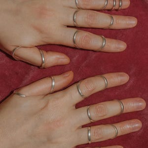 May include: Two hands adorned with numerous silver rings on each finger. The rings are slender and plain, offering a minimalist style. The backdrop is a deep red hue, contrasting with the silver jewellery.