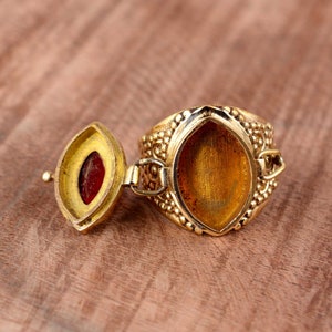 May include: A gold ring with a hinged lid that opens to reveal a small compartment. The ring is decorated with a textured pattern and has a smooth, oval-shaped interior.