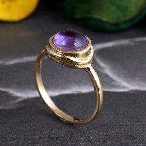 May include: A gold ring with a purple amethyst gemstone set in a bezel setting. The ring has a simple, elegant design.