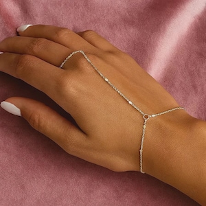 Puede incluir: Delicada pulsera de cadena de mano plateada con pequeños detalles redondos y ovalados. La pulsera se conecta a un anillo y se envuelve alrededor de la muñeca. La joya se exhibe sobre una superficie rosa.