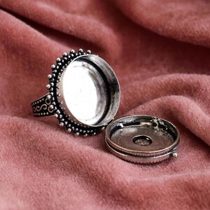 May include: A silver-colored ring with a large, round bezel setting and a decorative beaded edge. The ring is open, revealing the interior, and the matching lid is next to it. The ring sits on a pink fabric.
