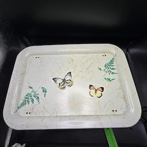 May include: A rectangular off-white serving tray featuring a pattern of scattered brown pine needles, two green fern sprigs, and two butterflies with yellow and black wings. The tray has four small metal rivets near the corners and a green ruler.