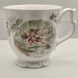 May include: A white ceramic mug with a scalloped base and a decorative handle, featuring a colorful illustration of a frog wearing a jacket and carrying a basket while fishing on a lily pad, surrounded by water lilies and green reeds.