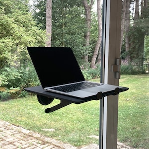 May include: A black laptop computer sits on a black shelf attached to a window. The laptop is open, displaying a black screen. The shelf is supported by a black bracket. The background shows a green yard and trees.
