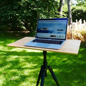 Puede incluir: Un ordenador portátil está sobre una mesa de madera clara sujeta a un trípode negro. La pantalla del portátil muestra un sitio web. La configuración está al aire libre, sobre un césped, con árboles al fondo.